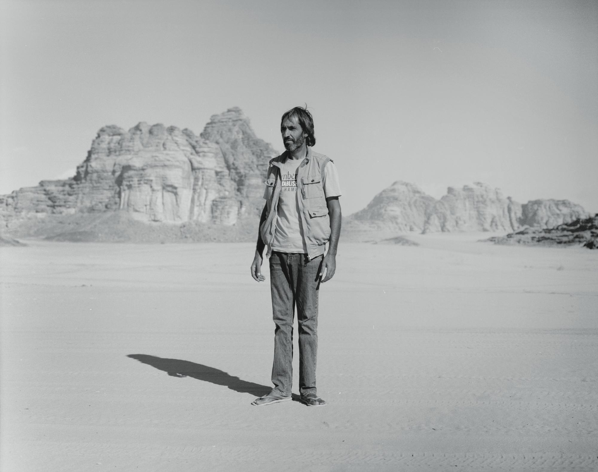 Monochrome outdoor portrait of a man in a vest and jeans standing in a desert landscape with rock formations in the background.