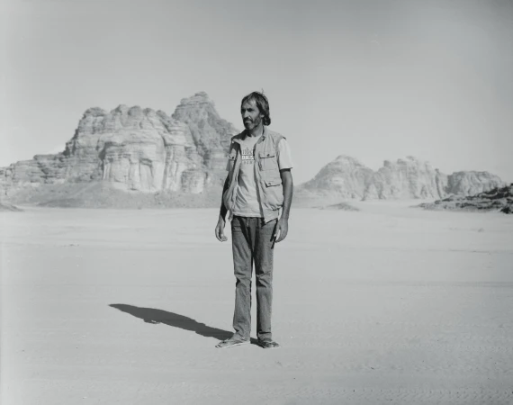 Monochrome outdoor portrait of a man in a vest and jeans standing in a desert landscape with rock formations in the background.