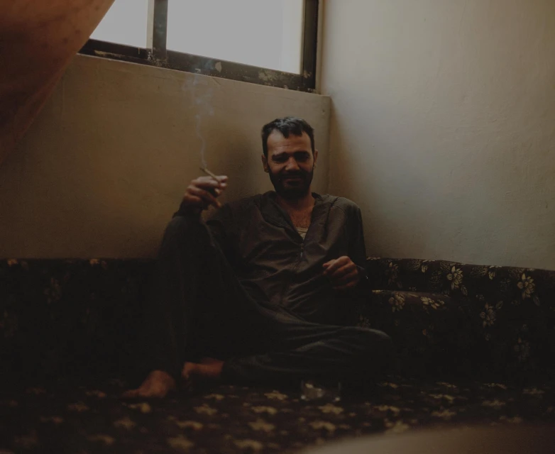Adult male in casual attire sitting indoors on patterned seating, holding a cigarette, with natural light from a nearby window.