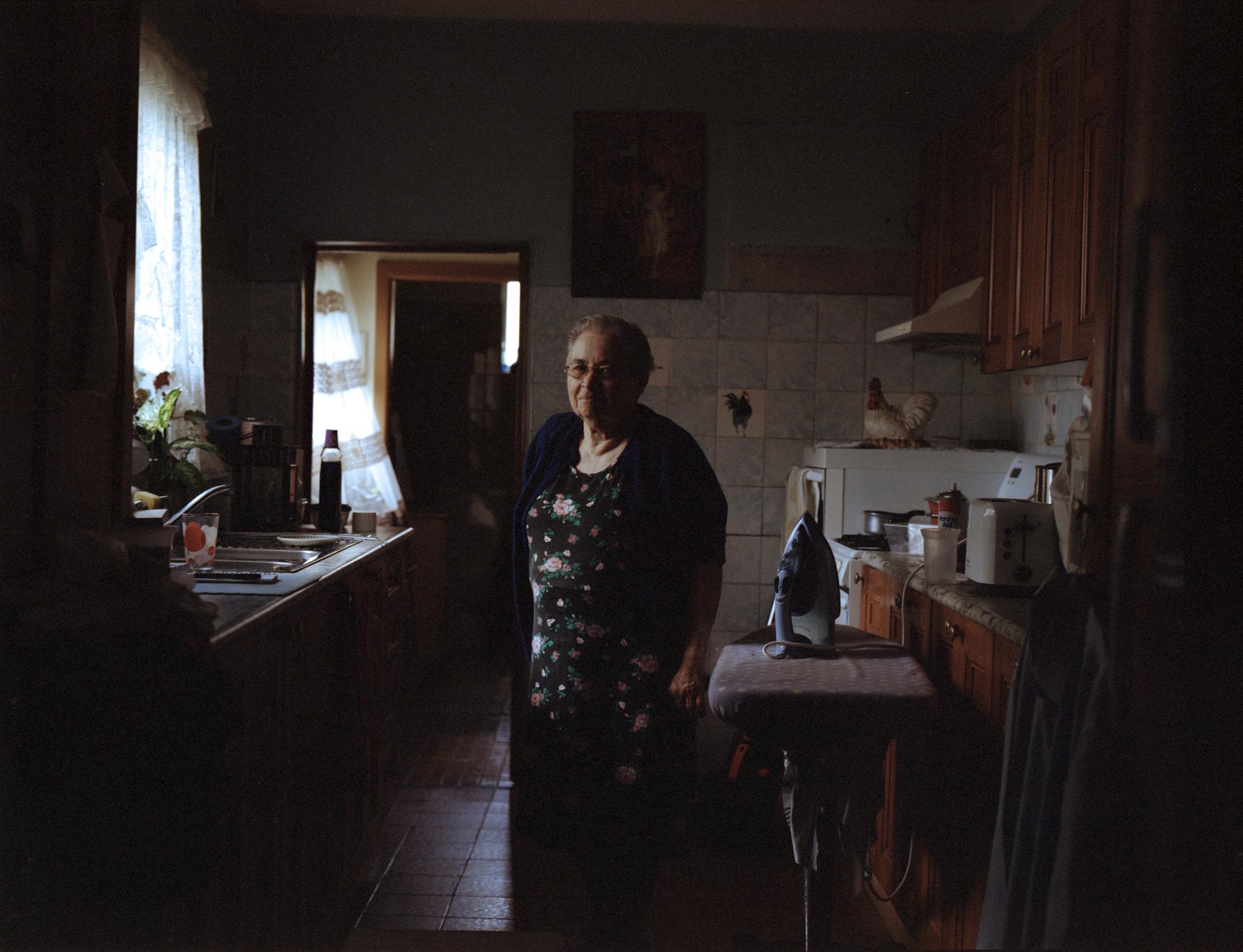 Indoor portrait of an elderly woman in a dress and cardigan, standing in a kitchen, with appliances and cabinetry in the background.