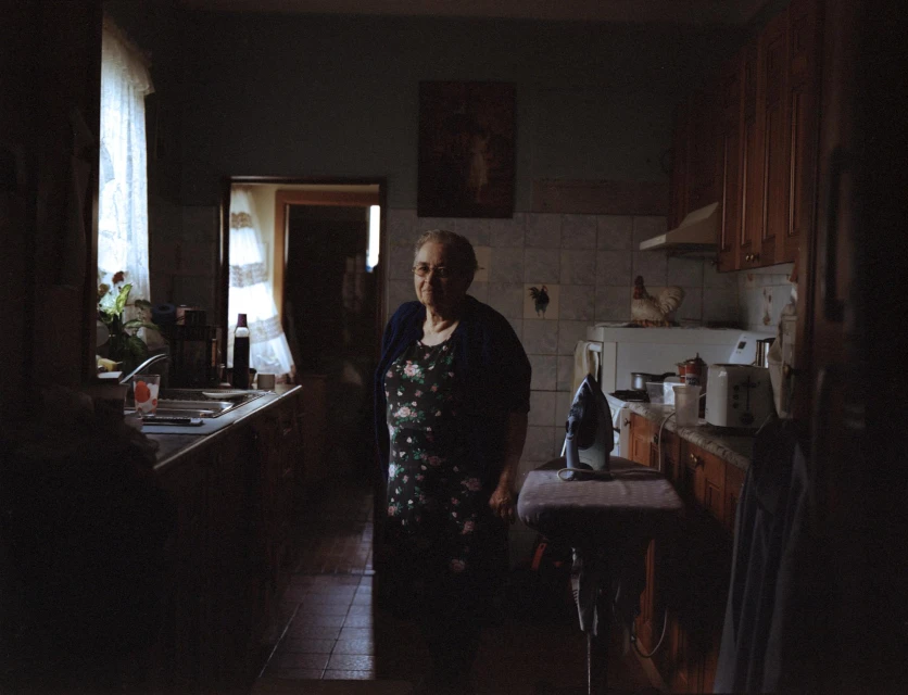 Indoor portrait of an elderly woman in a dress and cardigan, standing in a kitchen, with appliances and cabinetry in the background.