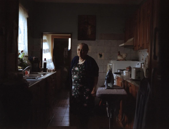 Indoor portrait of an elderly woman in a dress and cardigan, standing in a kitchen, with appliances and cabinetry in the background.