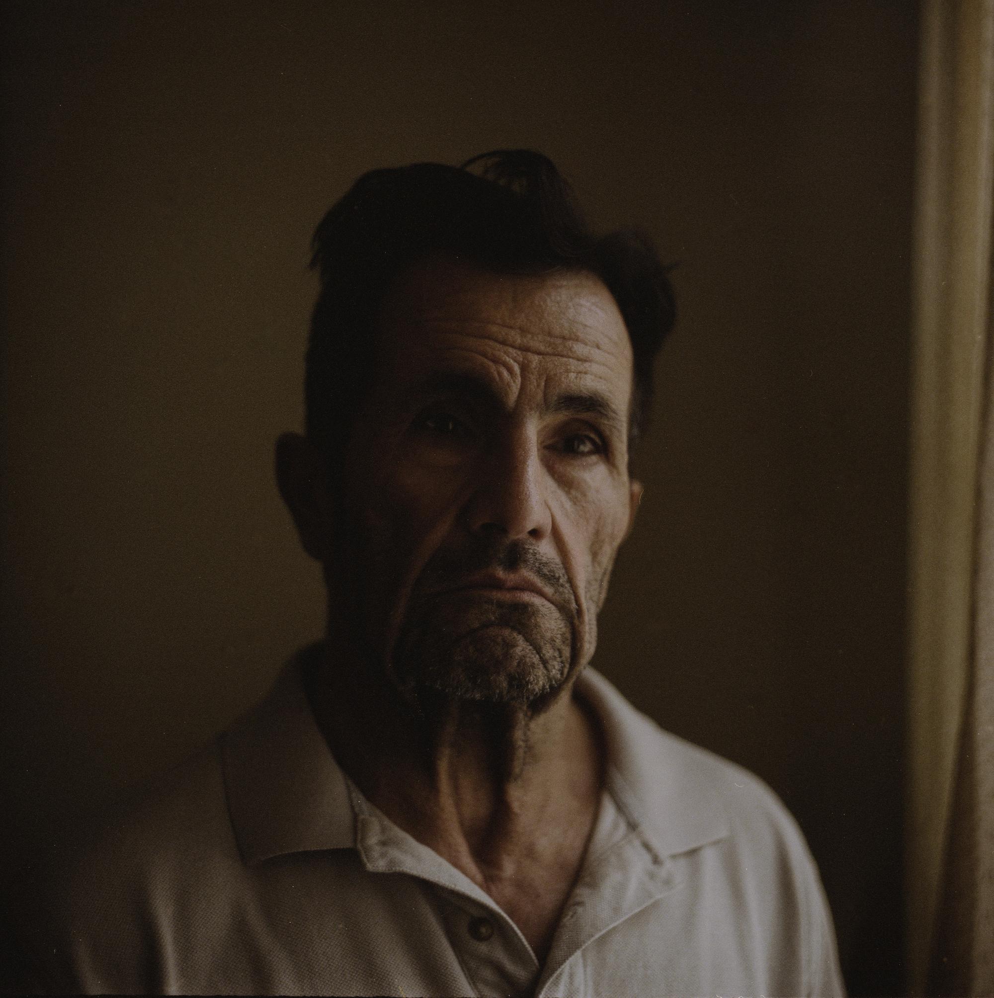 Close-up portrait of a mature adult male with dark hair and a white collared shirt against a dark grey background, medium shot.