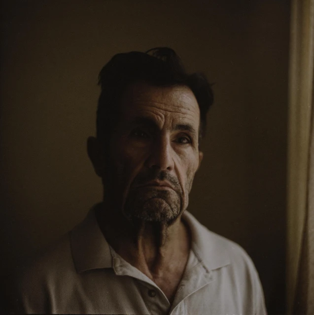 Close-up portrait of a mature adult male with dark hair and a white collared shirt against a dark grey background, medium shot.