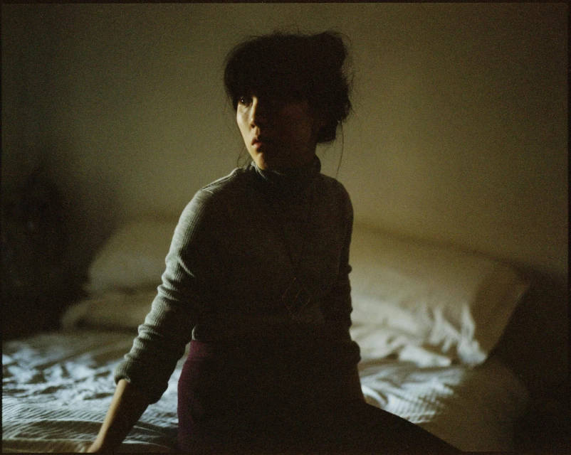 Indoor portrait of an adult woman sitting on a bed. She wears a grey sweater with a high neck, and the light is subdued.