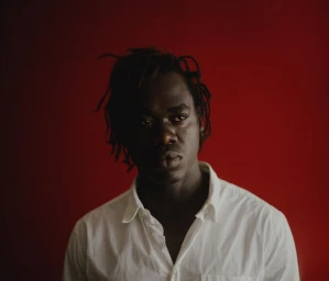 Portrait of a Black man with dreadlocks wearing a white button-down shirt against a dark red background by photographer Yeoseop Yoon