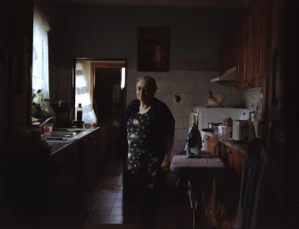 Elderly woman standing inside a dimly lit domestic kitchen with dark wooden cabinets and an ironing board