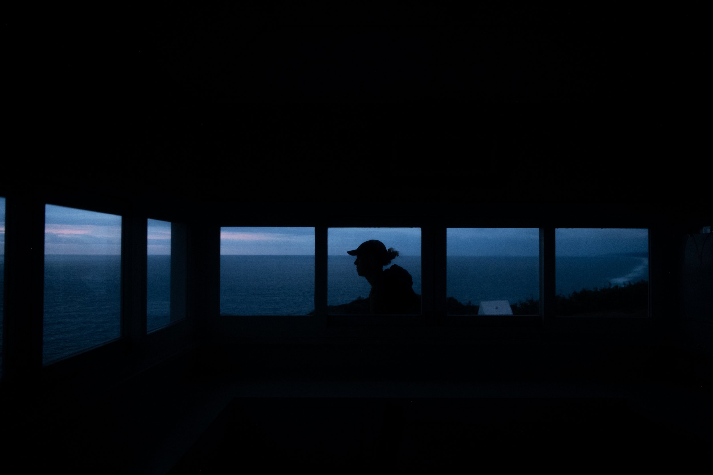 Silhouette of a person with a cap looking out at the sea through a window structure during nighttime, showing the horizon and ocean.