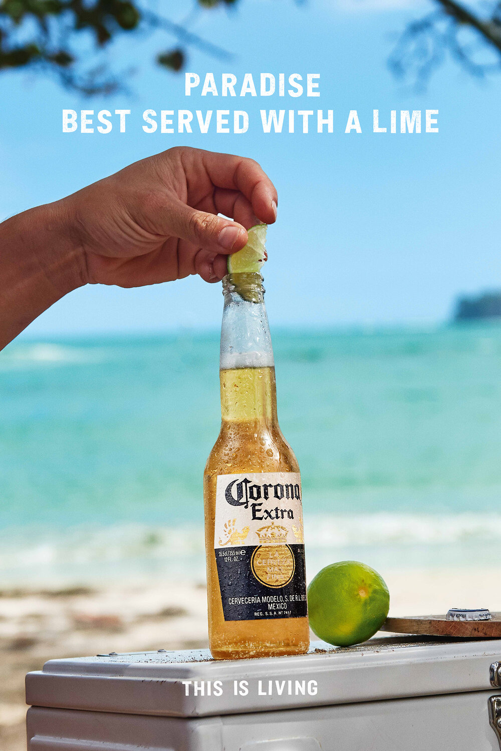 Person squeezing lime into a Corona beer bottle on a white surface with a beach and blue sky in the background. Refreshing beverage.