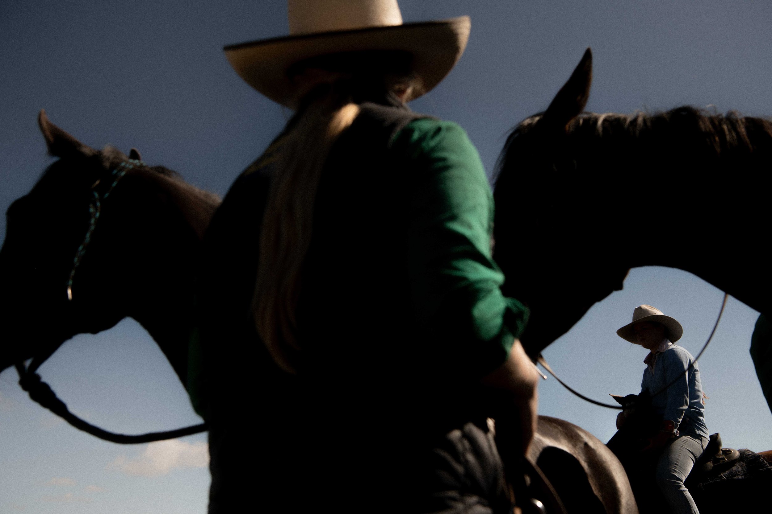 Two horses with riders in cowboy hats silhouetted against a blue sky, one rider in foreground, second rider in the background.