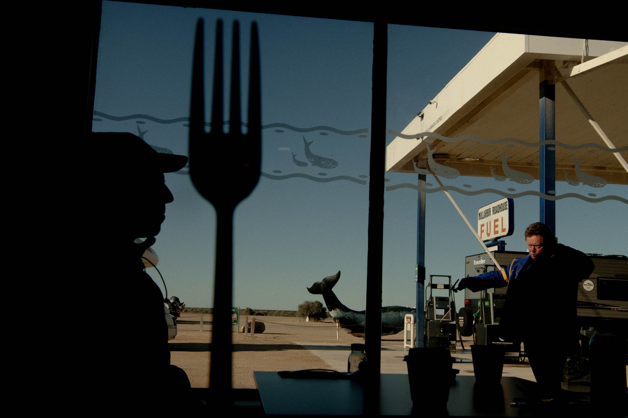 Silhouette of a man inside a diner with a giant fork silhouetted against a window view of a gas station and desert landscape.