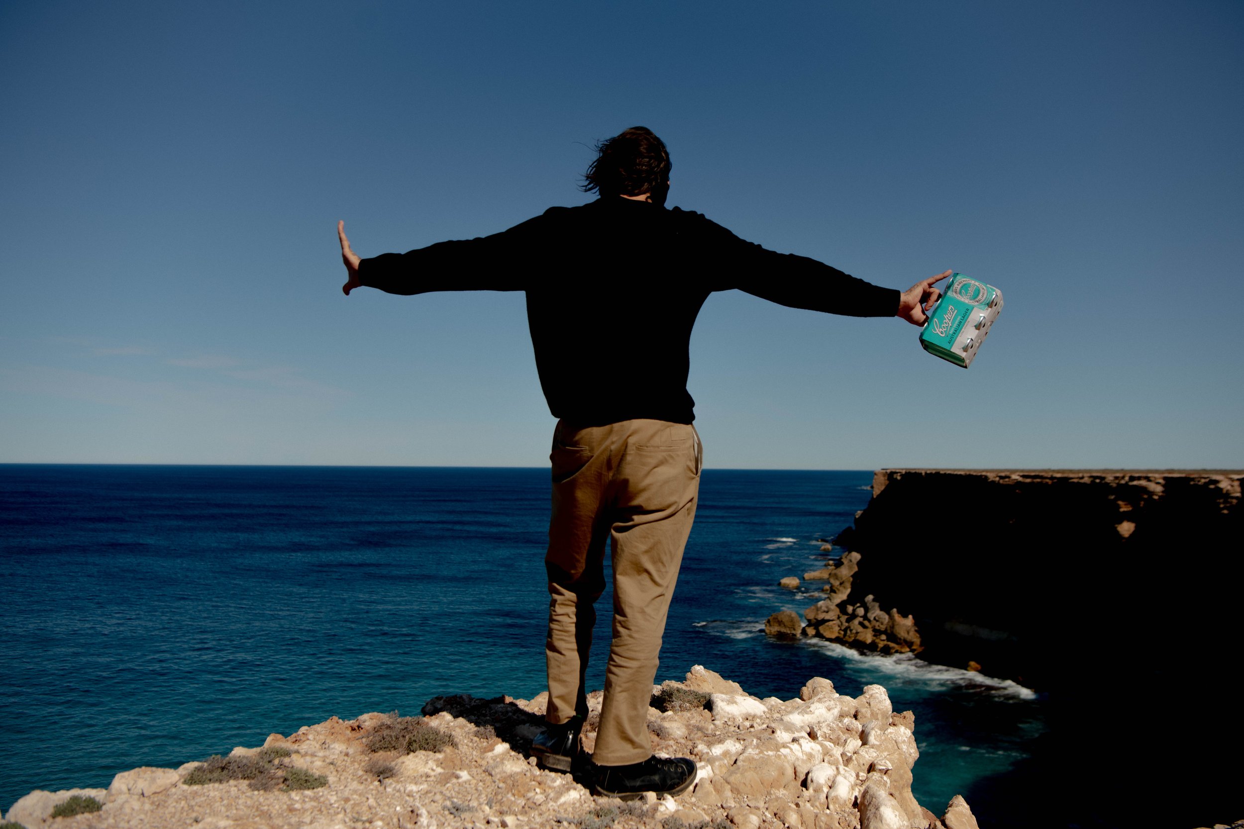 Adult stands on rocky cliff overlooking the ocean with arms outstretched and box in hand, under a clear blue sky on sunny day.