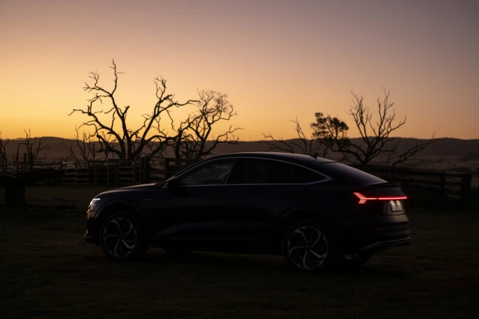 Dark Audi e-tron parked on a grassy field at dusk with bare trees and a fence in the background and an orange glow in the sky.