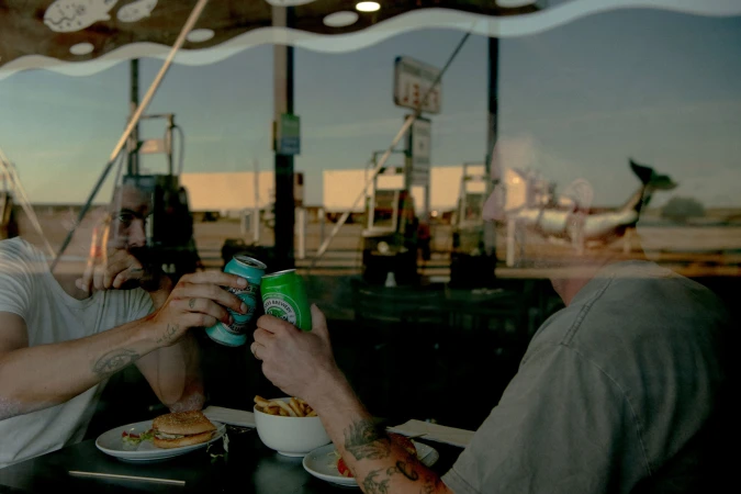 Two people toasting beer cans at a table with burger and fries. Gas station and passing cars reflected in window behind them.