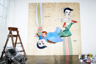 Woman suspended in front of large illustrated wooden panels featuring stylized figures, with a ladder and floor fan visible.