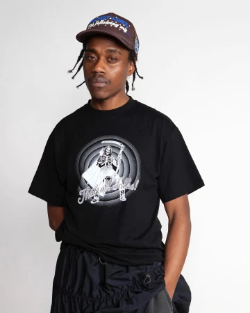 Black man wearing a black t-shirt and brown cap against a white backdrop photographed by Clare Shilland.