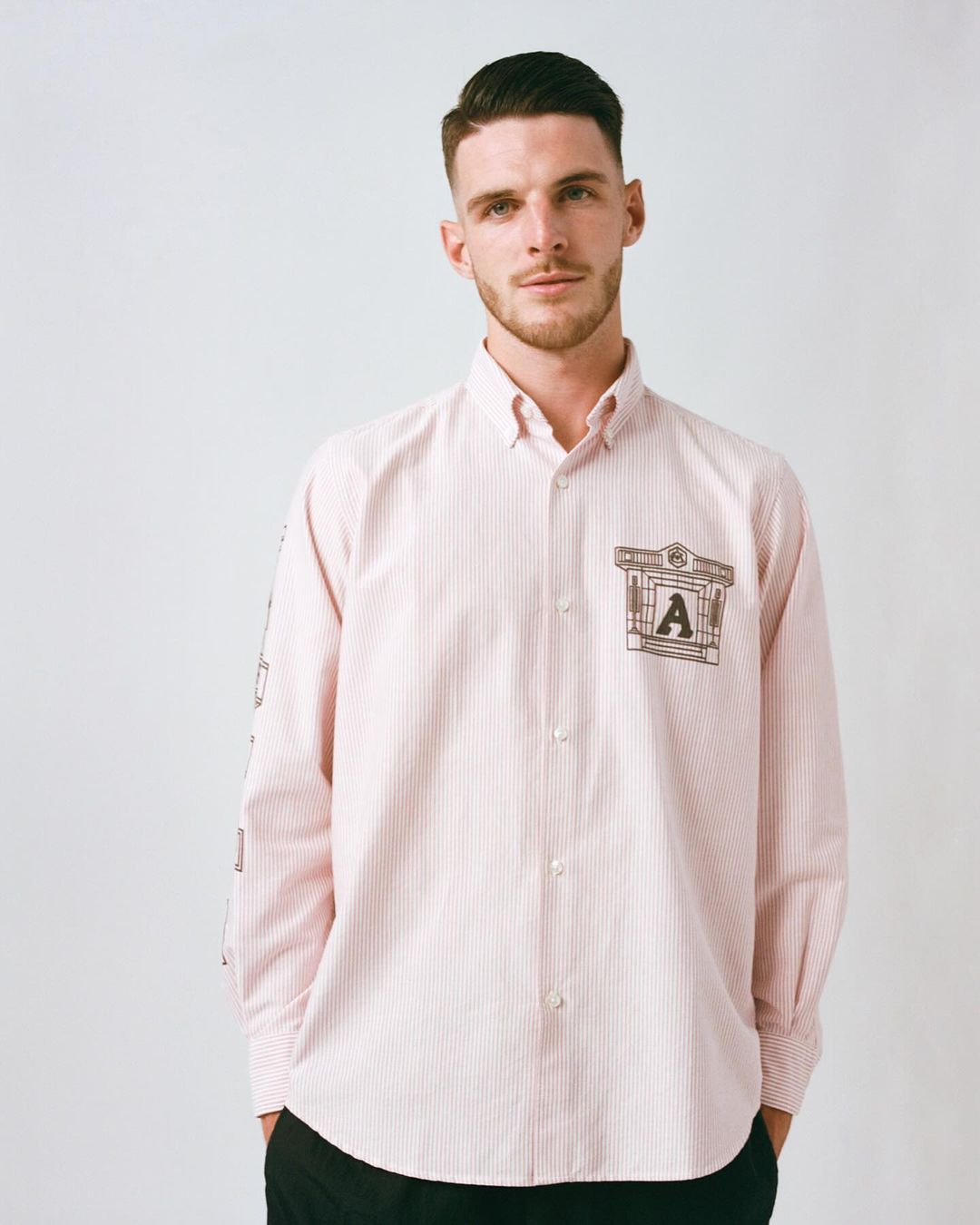 An adult male poses indoors, wearing a pink-and-white striped button-down shirt with a logo on the chest pocket and arm.