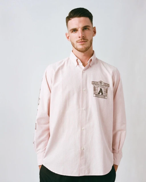 An adult male poses indoors, wearing a pink-and-white striped button-down shirt with a logo on the chest pocket and arm.