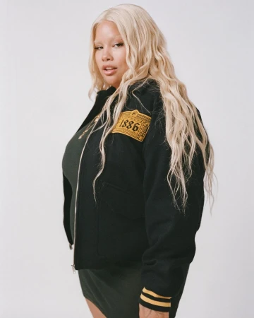 Blonde woman wearing a black jacket with yellow 1886 patch and dark green dress against a plain white backdrop