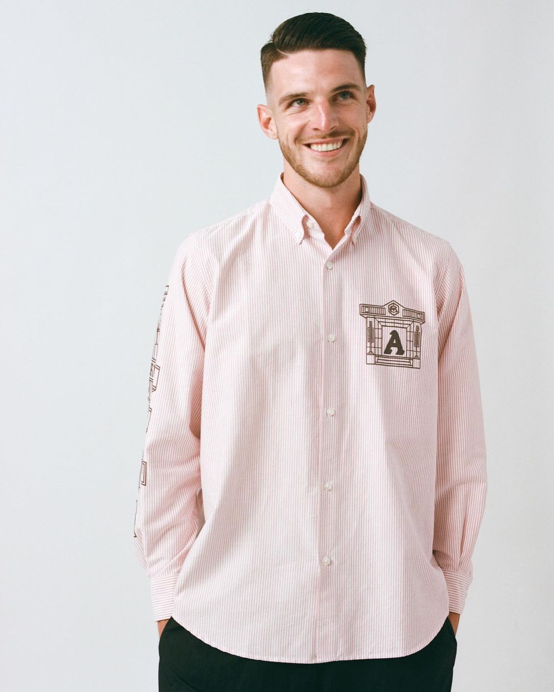 Handsome man in a light pink striped dress shirt with an architectural design and the letter 'A'. Smiling indoors against a white backdrop.