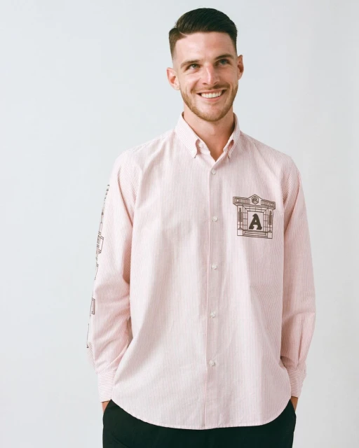 Handsome man in a light pink striped dress shirt with an architectural design and the letter 'A'. Smiling indoors against a white backdrop.