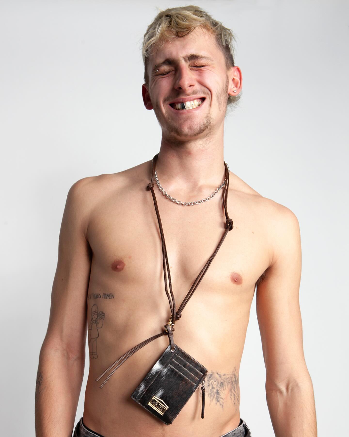 A topless adult man with blonde hair smiles and wears a black card holder with brown straps and a silver chain against a white background.