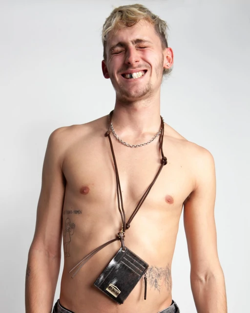 A topless adult man with blonde hair smiles and wears a black card holder with brown straps and a silver chain against a white background.