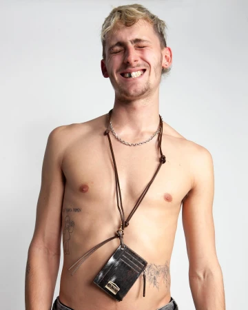 A topless adult man with blonde hair smiles and wears a black card holder with brown straps and a silver chain against a white background.