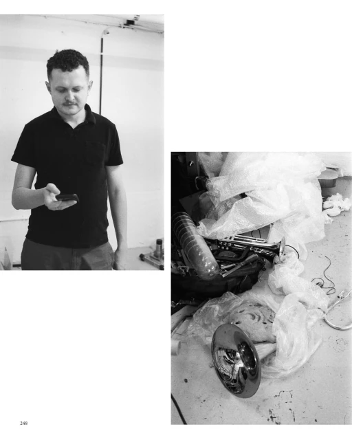 Monochrome image featuring an adult male looking at his smartphone on the left and a collection of objects piled on a surface on the right.