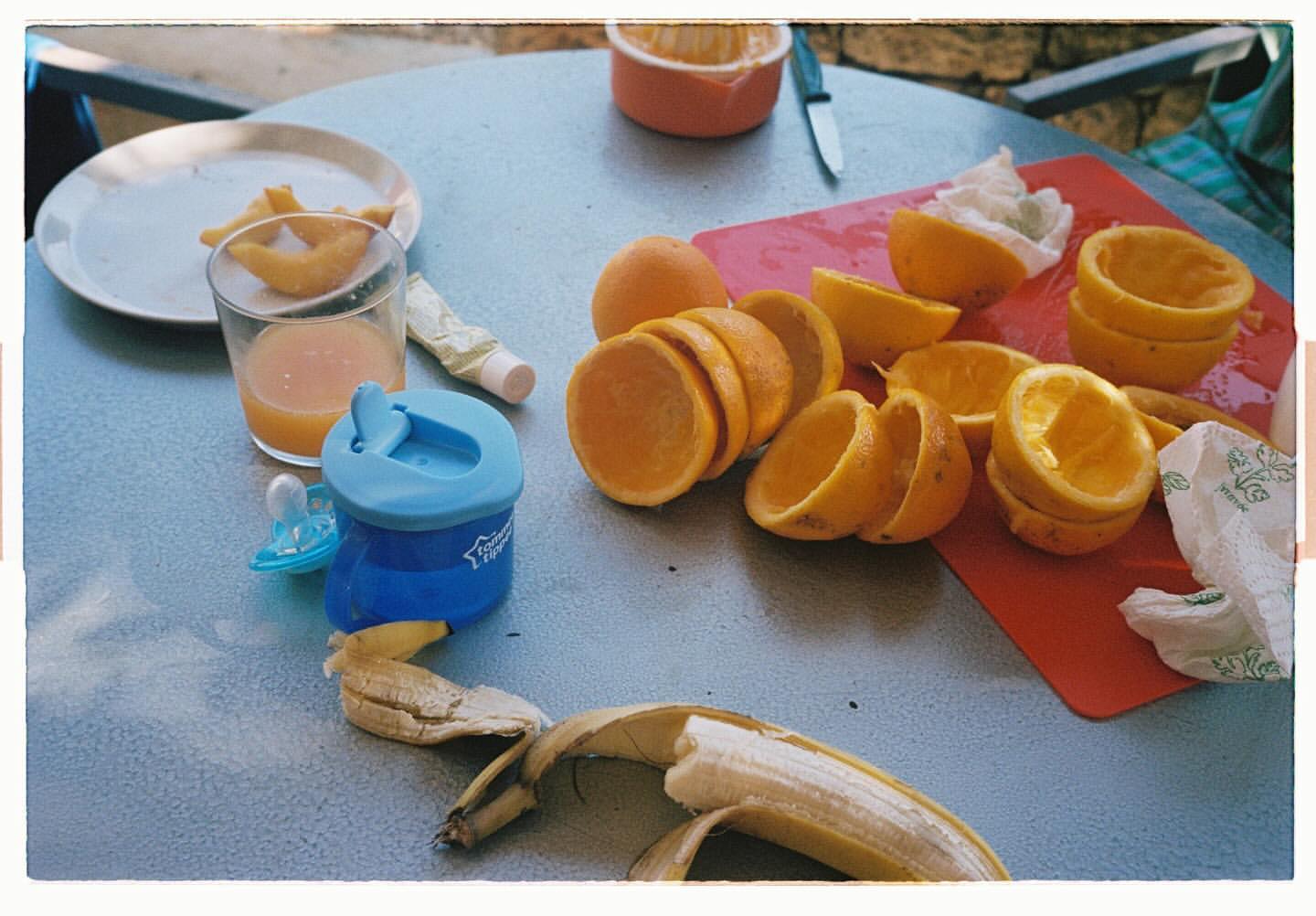 Tabletop still life of peeled oranges, banana peel, juice glass, baby bottle, and a cutting board, suggesting a healthy food preparation scene.
