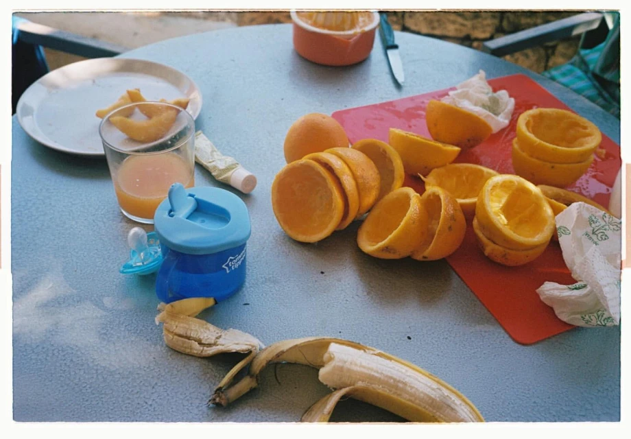 Tabletop still life of peeled oranges, banana peel, juice glass, baby bottle, and a cutting board, suggesting a healthy food preparation scene.
