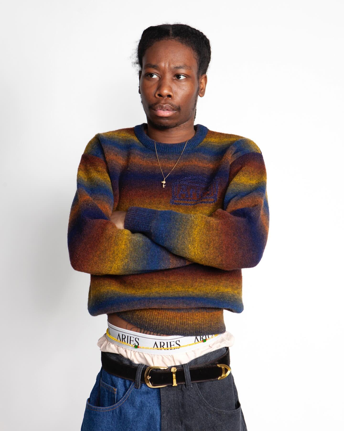 A young adult male poses in a studio wearing a striped sweater, partially visible underwear, jeans, and a belt against a white backdrop.