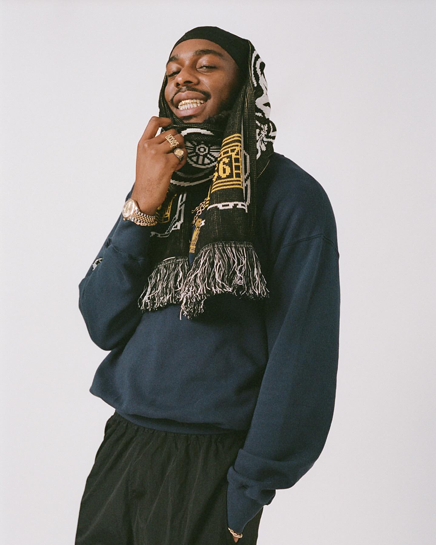 Portrait of a Black man wearing a navy sweatshirt, black pants, and a patterned black scarf wrapped around his head, photographed by Clare Shilland.