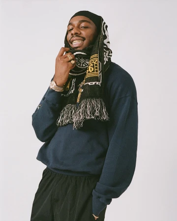 Portrait of a Black man wearing a navy sweatshirt, black pants, and a patterned black scarf wrapped around his head, photographed by Clare Shilland.