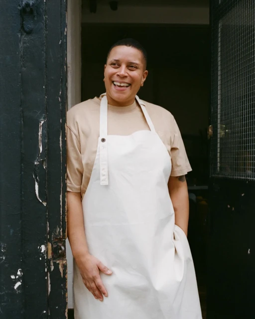 Smiling adult woman stands in a doorway wearing a tan T-shirt and white apron, bathed in soft light. Interior setting.