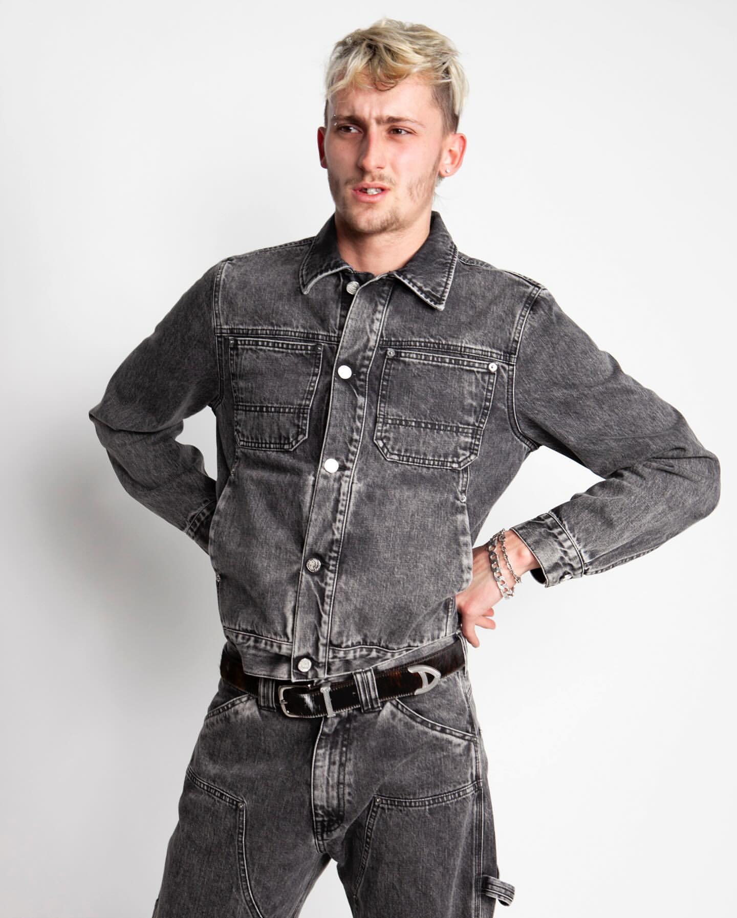Young adult man poses indoors wearing a grey denim jacket and matching pants, hands on hips, against a white backdrop.