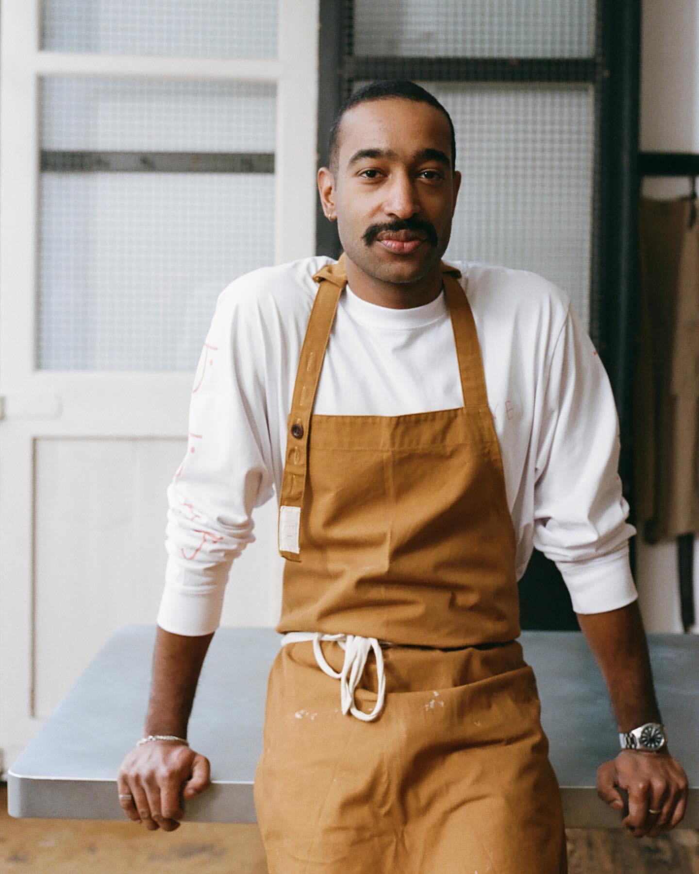 Portrait of an adult male with a mustache wearing a tan apron over a white long-sleeved shirt, standing indoors near a metal table.