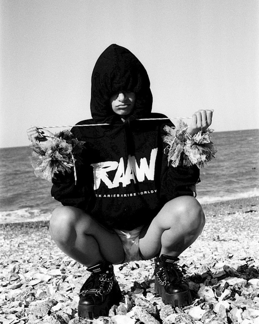 A monochrome medium shot shows a woman in a hooded sweatshirt crouching on a rocky beach with a line of textile art around her.