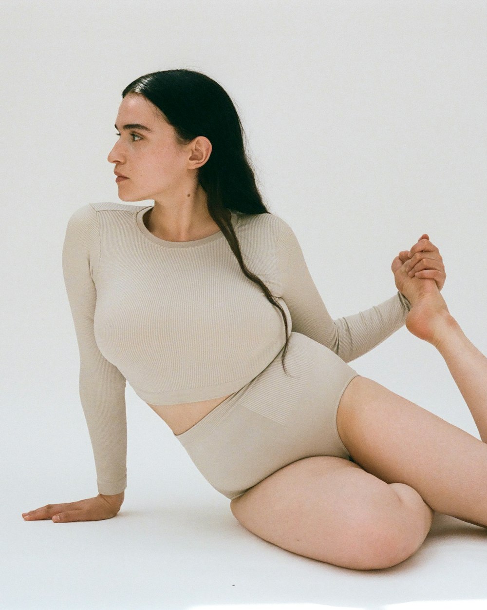 Woman in beige ribbed top and shorts poses on a white backdrop, looking away, showcasing a minimalist fashion and natural aesthetic.