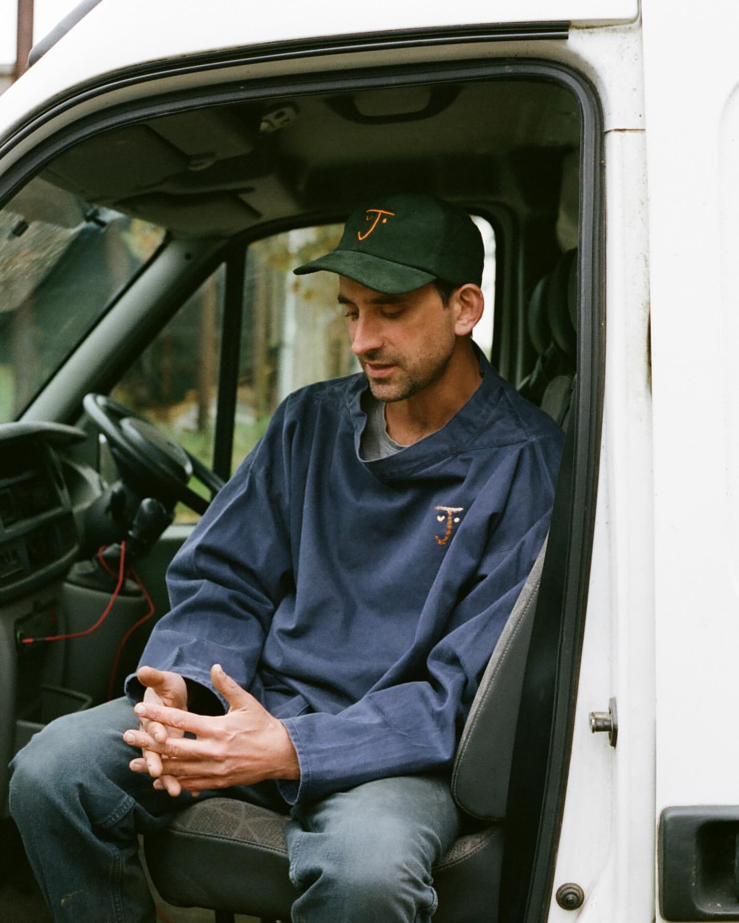 Man in a driver's seat wearing a cap and blue jacket with hands clasped in front of him. He sits inside a white delivery van.