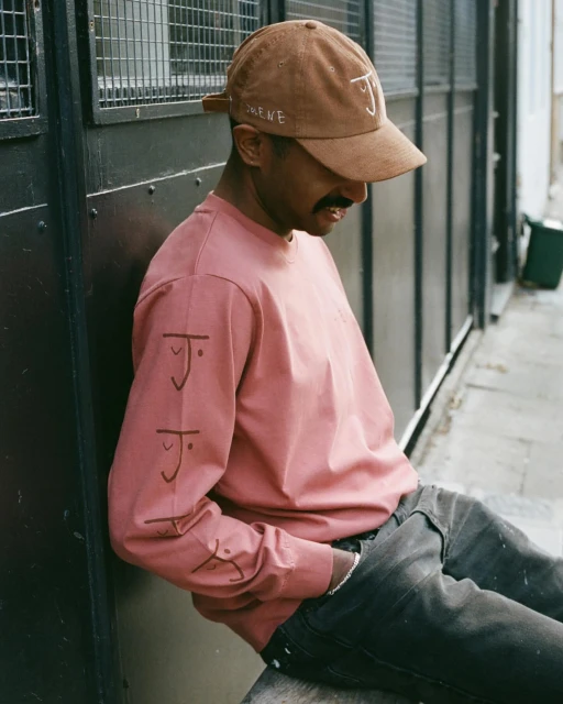 Man in a brown hat and pink shirt leans against a wall in an urban setting, showcasing casual street style and neutral tones.