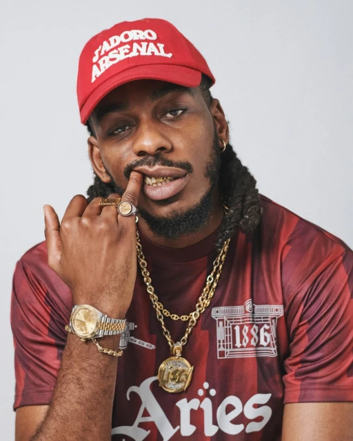 Adult male with dreadlocks poses in front of a white wall, wearing a baseball cap, gold jewelry, and a sports jersey.