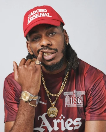 Adult male with dreadlocks poses in front of a white wall, wearing a baseball cap, gold jewelry, and a sports jersey.