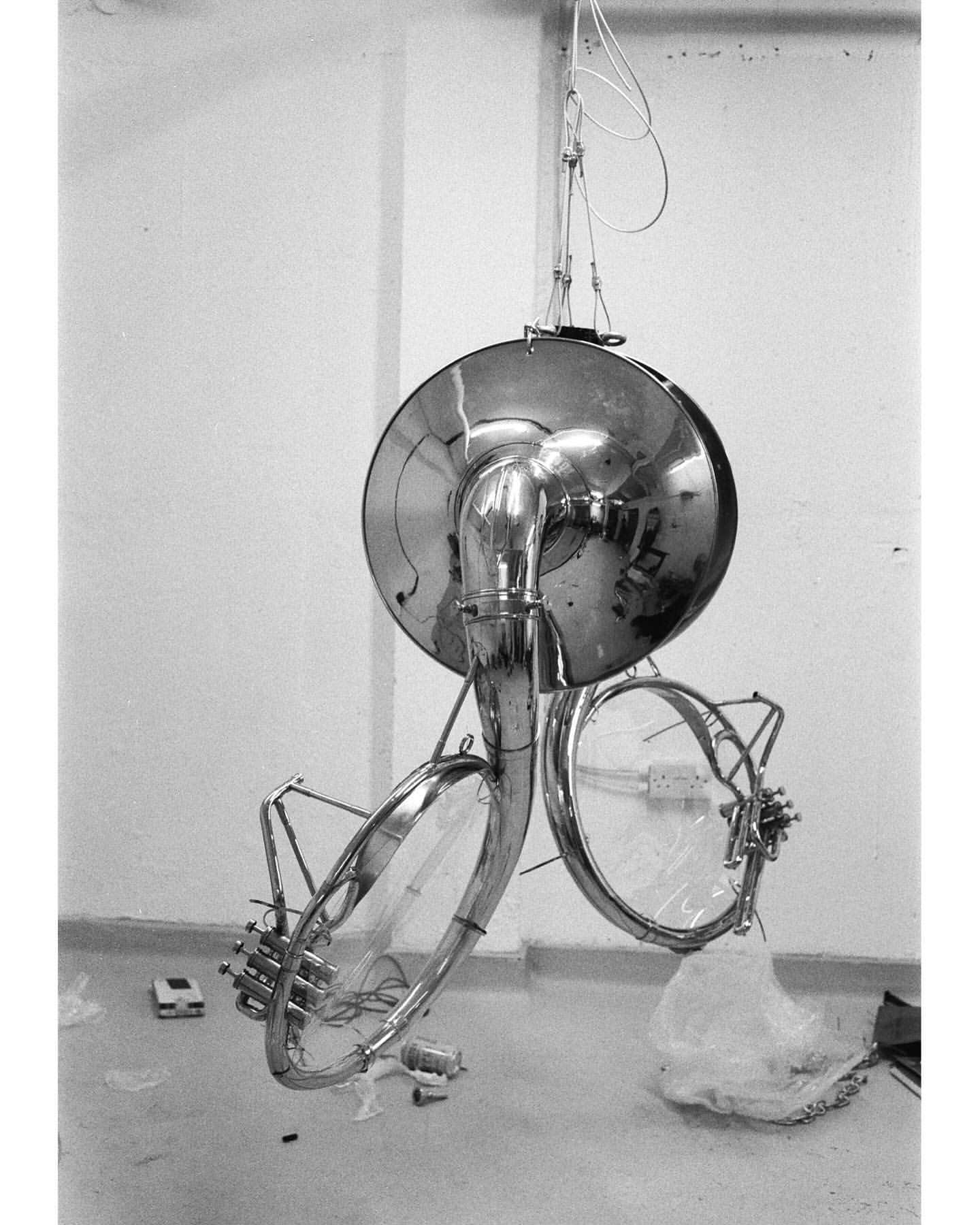 Black and white photograph of a disassembled sousaphone hanging from wires in an indoor setting with white walls and a gray floor.
