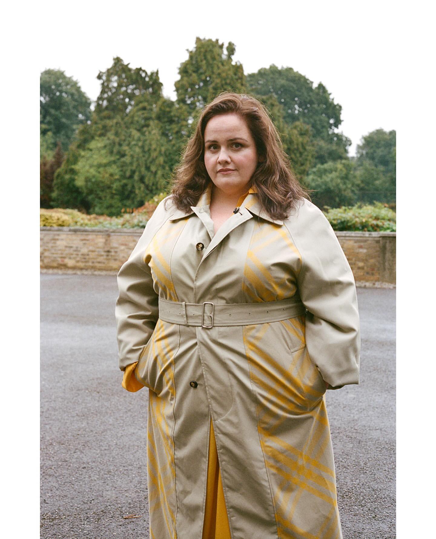 An adult woman is posing outdoors in a tan trench coat with yellow accents, set against a blurred green background with trees and sky.