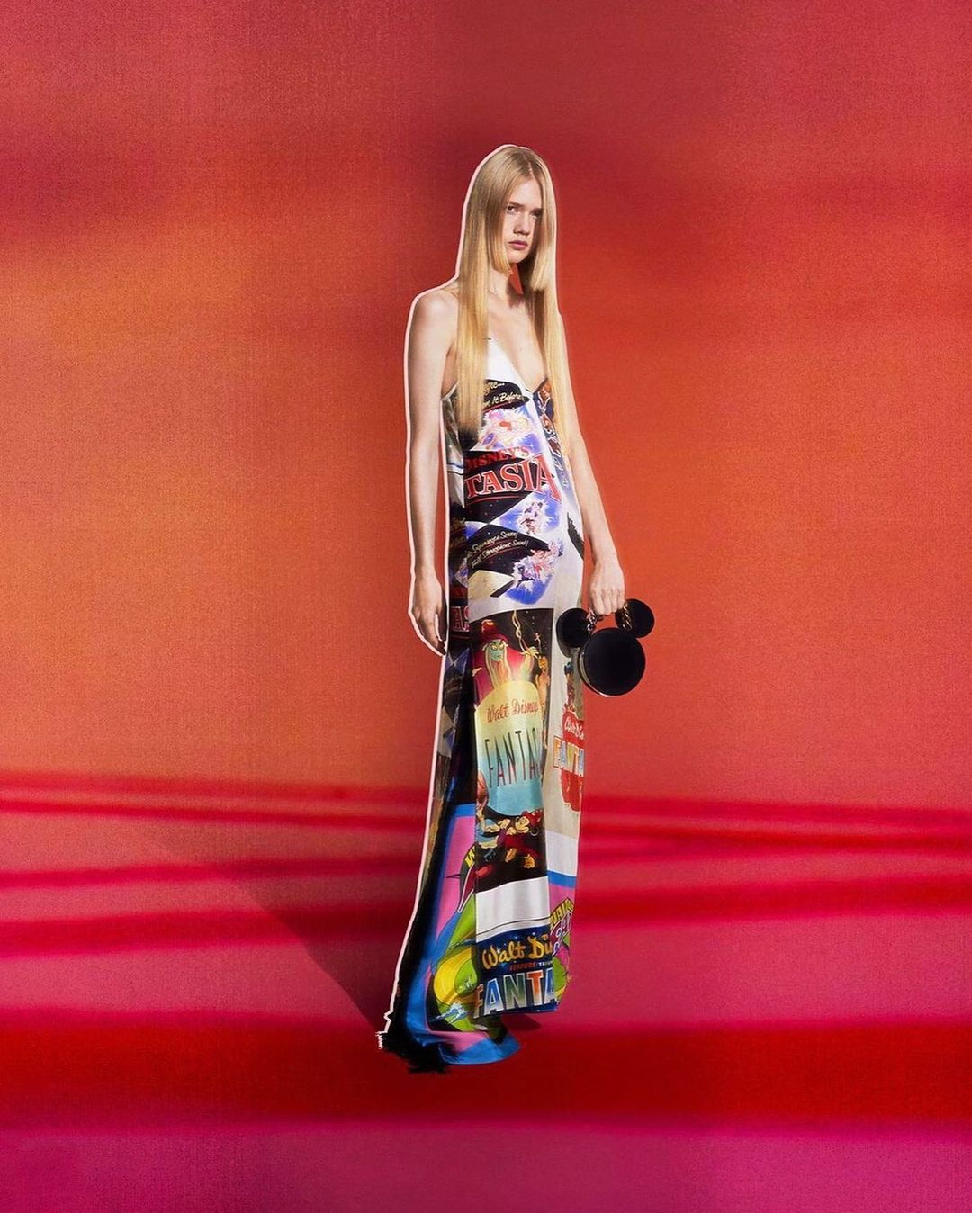 Fashionable woman in a printed maxi dress holding a Mickey Mouse purse, posing in front of a vibrant orange and red backdrop.