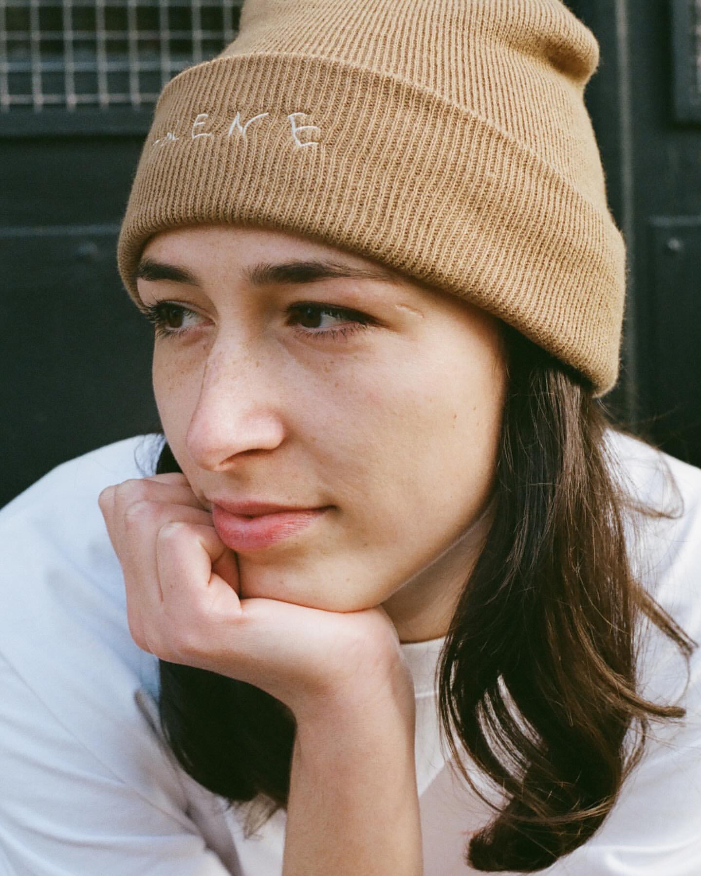 Close-up portrait of a young adult woman with long brown hair and a tan beanie cap, looking relaxed in an urban outdoor setting.