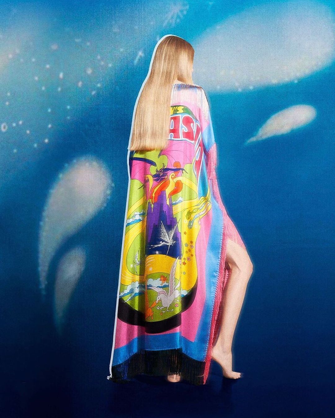 Adult female wearing a colorful dress with an illustration stands in front of a blue background with soft white shapes. Fashion shot.