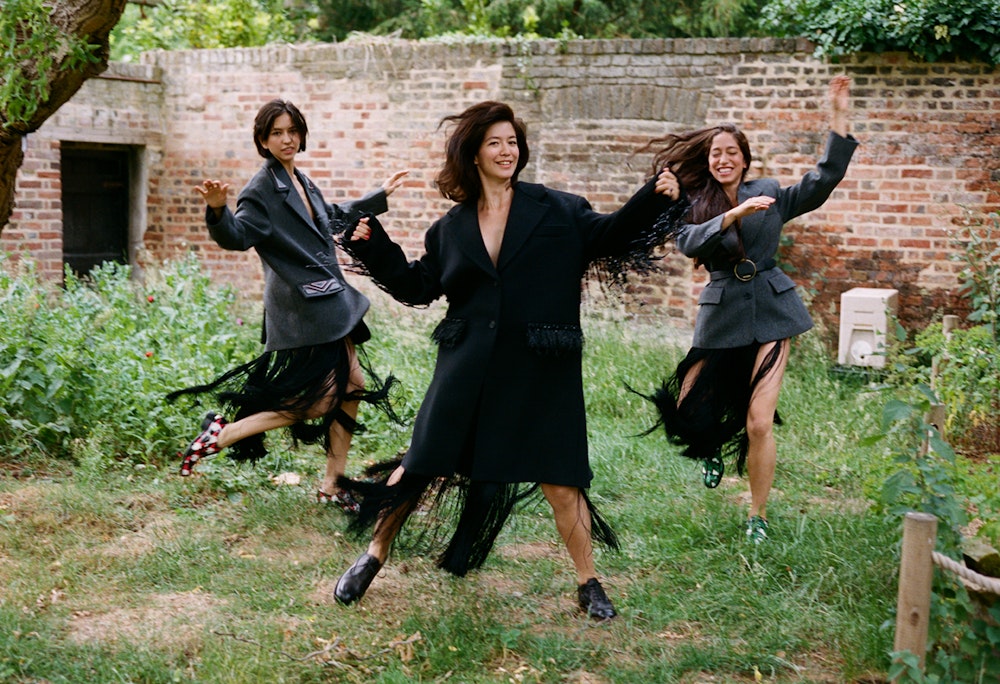 Three women in fashionable coats with fringe details jump joyfully in a grassy yard with a brick building in the background on a sunny day.