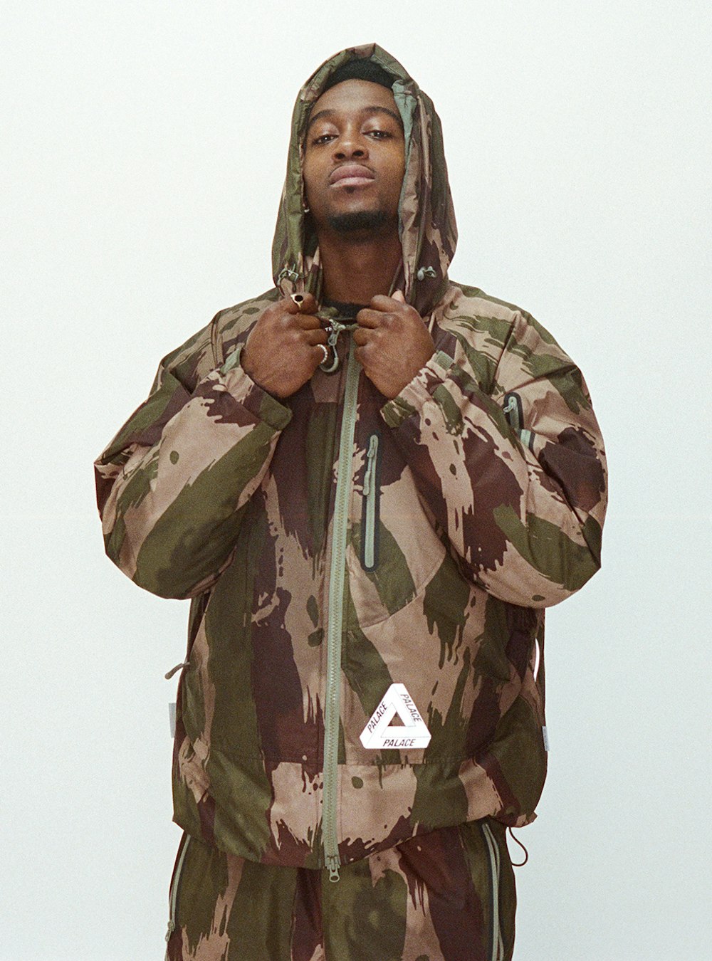 A Black male model in a camouflage jacket with a hood stands indoors against a white backdrop, showcasing modern streetwear fashion.